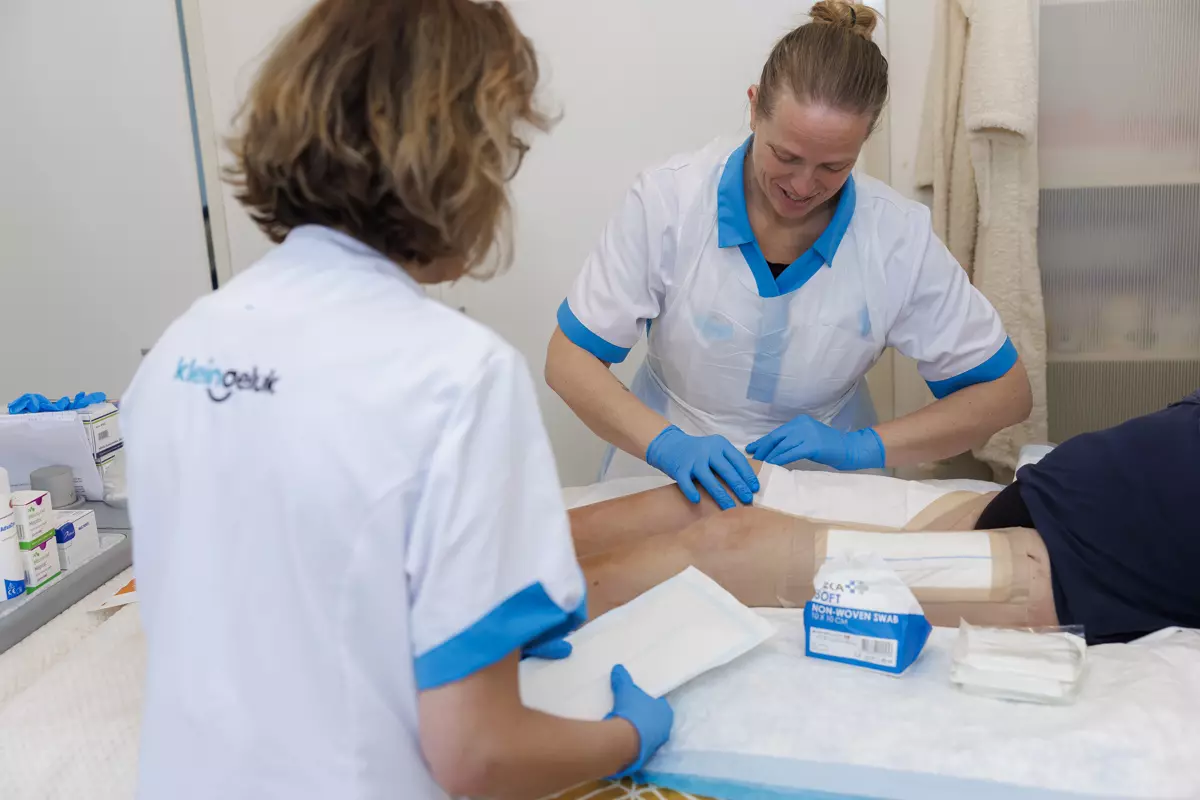 Twee verpleegkundigen van KleinGeluk verlenen wondzorg aan de bovenbenen van een oudere.
