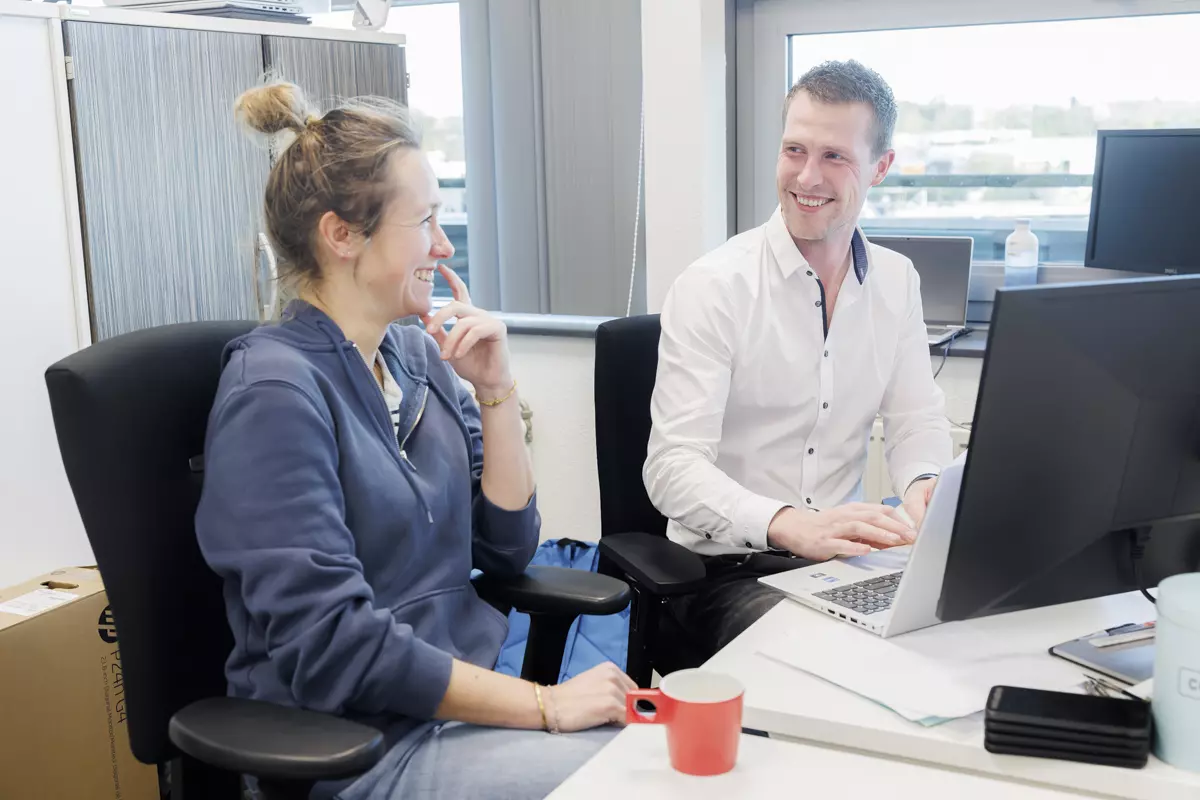 ICT medewerker van KleinGeluk legt een collega wat uit. Ze zitten samen achter een bureau en lachen om een grap.