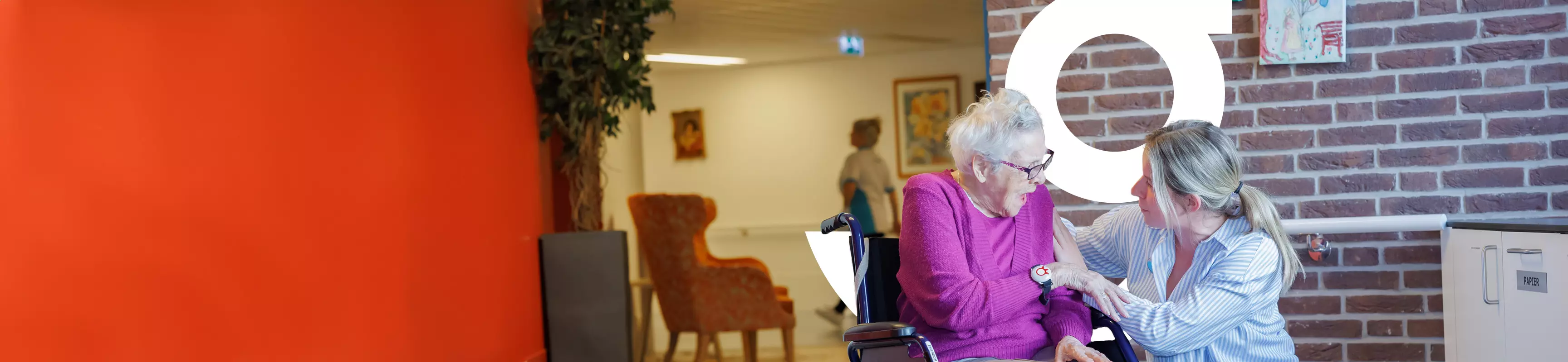 Een oudere dame zit in een rolstoel. Een verzorgster zit naast haar op haar hurken. Ze praten met elkaar
