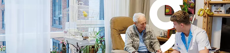 Een oudere man zittend in een stoel heeft een geanimeerd gespreek met een zorgmedewerker van KleinGeluk. Beeld van zorg en comfort.