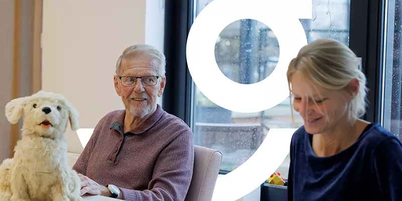 Een oudere en een woonassistent  van KleinGelukhebben een onderonsje over de knuffelhond op de tafel voor ze
