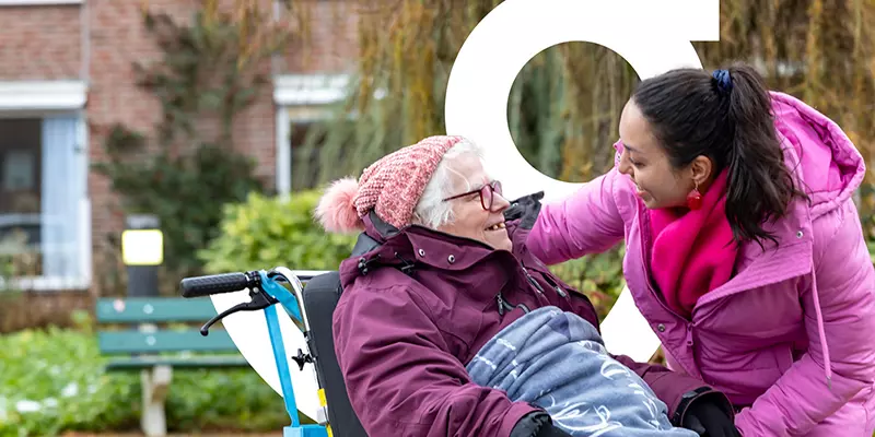 Een vrijwilligster heeft een onderonsje met een oudere mevrouw in een rolstoel terwijl ze lekker buiten zijn.